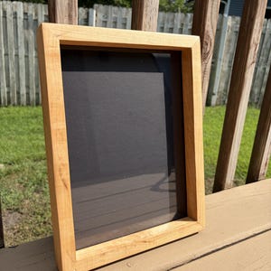 Puede incluir: Un marco de fotos rectangular construido con madera natural de tono claro con un acabado liso. El marco presenta un diseño de caja profunda y contiene un panel de fondo negro sólido colocado dentro del centro del borde de madera.