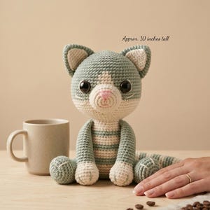 Pode incluir: Boneca de gato de crochê, com aproximadamente 25 cm de altura, em tons de cinza, branco e rosa. O gato feito à mão tem olhos grandes e pretos e está sentado em uma superfície de madeira. À esquerda do gato há uma caneca bege.