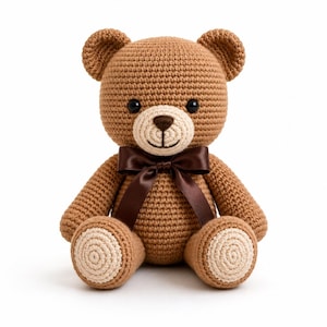 Puede incluir: Un oso de peluche tejido a ganchillo en color marrón con un hocico color crema y parches circulares a juego en las plantas de los pies. El oso tiene ojos de plástico negro, nariz y boca bordadas en marrón oscuro y un lazo de satén.