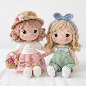 Puede incluir: Dos muñecas de ganchillo hechas a mano. Una muñeca lleva un sombrero de paja con flores, una blusa blanca y una falda rosa con bordados florales. La otra tiene una diadema azul y un pelele verde. Ambas muñecas tienen ojos negros y mejillas rosadas.