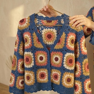 May include: A blue crochet cardigan with a V-neck and long sleeves. The cardigan features a floral pattern in shades of pink, yellow, and brown. The garment is displayed on a gold hanger.