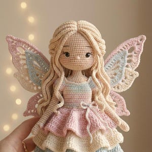 May include: A handmade crochet fairy doll with long blonde hair and delicate features. The doll wears a pastel-colored dress with a layered skirt and matching wings in shades of pink, blue, and cream. The doll is approximately 10 inches tall.