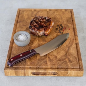 Pode incluir: Uma tábua de corte retangular de madeira de topo com ranhura para sucos e recorte para pega. Na tábua encontram-se um bife selado, uma pequena taça de betão cinzento com sal marinho grosso, uma faca de chef com cabo vermelho escuro e grãos de pimenta.