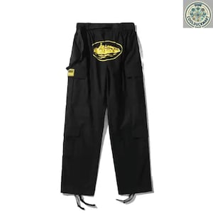 May include: Black cargo pants with a yellow oval design on the back. The pants have multiple pockets, a belt, and drawstrings at the ankles. A small yellow tag is visible on the left side. The pants are displayed on a white background.