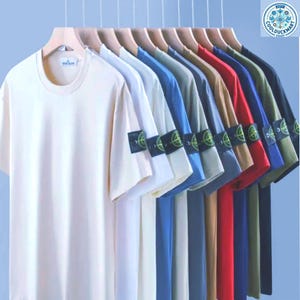 Puede incluir: Una colección de camisetas de manga corta en varios colores, incluyendo crema, blanco, azul y rojo, colgadas en perchas de madera. Cada camiseta tiene un pequeño parche en la manga izquierda. Las camisetas se muestran sobre un fondo azul claro.
