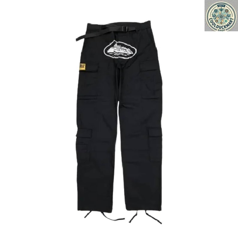 May include: Black cargo pants with multiple pockets, a black belt, and a white and black graphic on the front. The pants have drawstrings at the ankles and a small yellow patch. The pants are displayed on a white background.