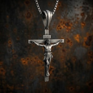 Puede incluir: Un colgante de crucifijo de metal en tono plateado oscuro que presenta una figura detallada sobre una cruz con las letras I.N.R.I. inscritas sobre la figura. El colgante cuelga de una cadena de caja de metal plateado oscuro a juego.