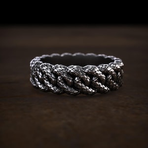 Könnte beinhalten: Ein dunkel silberfarbener Ring mit einem durchgehenden Kettenglied-Design. Die Metalloberfläche ist mit einem detaillierten, überlappenden Schuppenmuster texturiert, das jedes einzelne Glied bedeckt und ein einheitliches, komplexes Erscheinungsbild erzeugt.