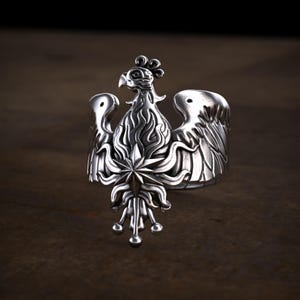 May include: A silver-toned ring featuring a detailed bird design with spread wings, a crest on its head, and a prominent star-shaped emblem at the center of its chest, with decorative flame-like patterns extending downward from the star.