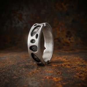 Könnte beinhalten: Ein silberfarbener Ring mit einem breiten Band und einem organischen Ausschnittmuster, das ein dunkles, vertieftes Inneres offenbart. Die Außenfläche hat ein gebürstetes, mattes Finish mit unregelmäßigen, abgerundeten Öffnungen, die das dunklere Metall freilegen.
