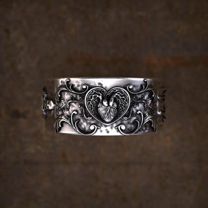 May include: A wide silver ring featuring a central anatomical heart motif enclosed within a heart-shaped outline. The band is decorated with intricate, raised scrollwork and filigree patterns that extend across the polished metallic surface.