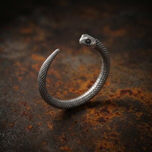 May include: A silver-toned metal cuff bracelet shaped like a coiled snake. The surface of the bracelet features a detailed, textured scale pattern throughout the entire length, ending in a sculpted snake head with defined eyes and facial features.