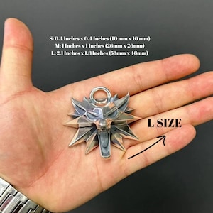 May include: A silver-colored wolf head pendant with a metallic finish and a top loop. Text indicates sizes: S: 0.4 inches x 0.4 inches, M: 1 inch x 1 inch, and L: 2.1 inches x 1.8 inches. An arrow points to the pendant labeled as L SIZE.