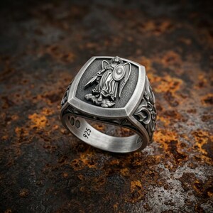 Puede incluir: Un anillo de plata de ley con una cara rectangular que presenta un relieve de una figura alada sosteniendo una espada y un escudo. El interior de la banda está claramente marcado con el número 925, indicando la pureza de la plata del metal.