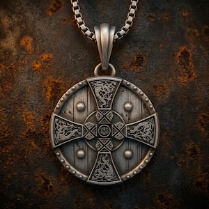 May include: A silver-toned circular pendant on a box chain, designed to resemble a wooden shield with a central cross. The pendant features intricate embossed knotwork, dragon-like motifs, and small circular studs along the outer rim.