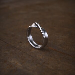Op de afbeelding: Een zilverkleurige ring met een gladde, doorlopende band met een enkele elegante draai aan de bovenkant. Het metaal heeft een zachte, matte afwerking die het licht opvangt langs de gebogen randen, wat een strakke vorm creëert.