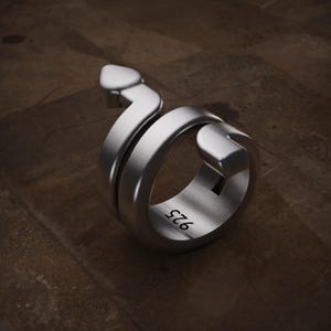 Puede incluir: Un anillo de metal color plata con forma de serpiente enroscada con una cabeza puntiaguda y un extremo de cola plano. La banda interior del anillo presenta el texto grabado 925, que indica la composición de plata esterlina de esta joya.