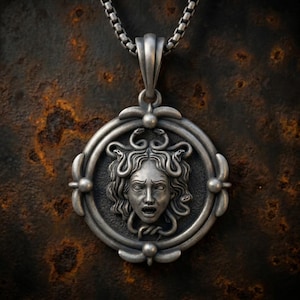 May include: A circular silver-toned pendant featuring a detailed relief of a face with snakes for hair, suspended from a matching box chain. The pendant has a dark, oxidized finish and a decorative frame with four small spherical accents at the edges.