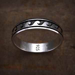 May include: A silver band ring featuring a continuous embossed wave pattern set against a dark, oxidized background. The interior of the band is clearly stamped with the number 925, indicating the sterling silver composition of the jewelry piece.