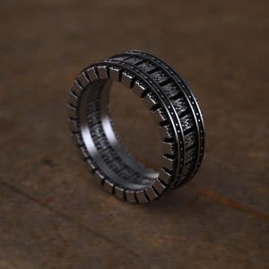 May include: A dark silver-toned metal ring featuring a wide band with a textured, gear-like outer edge. The surface displays a repeating pattern of rectangular segments separated by thin grooves, creating a mechanical appearance with a matte finish.