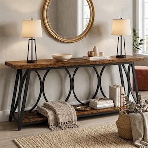 May include: A long console table with a dark wood grain top and a lower shelf, supported by a black metal frame featuring circular accents. Two black table lamps with beige shades sit on the top surface alongside a bowl and a book.