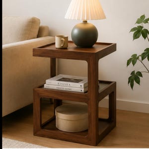 May include: A dark wood side table with two shelves. The top shelf holds a lamp with a cream-colored shade, a mug, and a dark green lamp base. The lower shelf has a round box and two books that read "KINFOLK" and "THIS IS HOME".