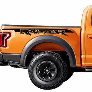 May include: Orange pickup truck with black "RAPTOR" decal on the side. The truck has black tires and a black fender. The decal is black with gold lettering. The truck bed has a black stripe.