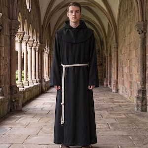 May include: A floor-length black robe featuring long sleeves, a hood, and a shoulder capelet. The garment is cinched at the waist with a thick, off-white rope belt that hangs down the front, tied in a simple knot. The fabric appears solid and opaque.