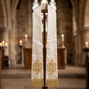 May include: A cream-colored liturgical stole featuring gold-colored embroidered Celtic knot designs at the base of each panel. The bottom edges are finished with gold-colored fringe trim. The stole is draped over a wooden stand.
