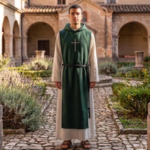 May include: A long-sleeved white robe worn under a dark green hooded tabard featuring a gold cross emblem on the chest. A black braided rope belt is tied around the waist, securing the tabard over the robe. The garment reaches the ankles.