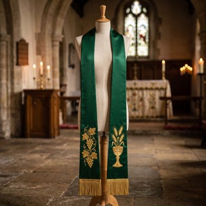 May include: A dark green liturgical stole featuring gold embroidery on both ends. One side displays a cluster of grapes with leaves, while the other side shows a chalice with wheat stalks. The bottom edges are finished with gold fringe trim.