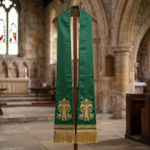 May include: A green liturgical stole featuring gold embroidered Tau crosses, stylized floral motifs, and two birds at the base of each panel. The stole is finished with gold fringe at the bottom edges and is displayed on a wooden stand.