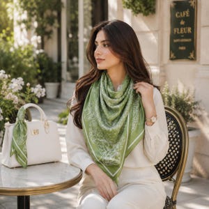Pode incluir: Uma mulher usando um lenço de seda verde claro com um desenho floral branco. Um lenço combinando está amarrado em uma bolsa branca com detalhes dourados. A mulher está sentada em uma mesa ao ar livre.