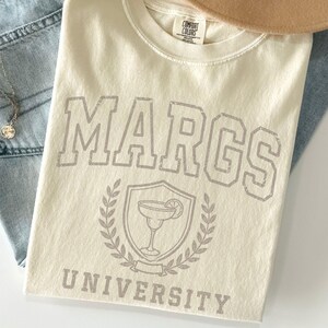 May include: A cream-colored short-sleeve t-shirt featuring the text MARGS UNIVERSITY in a collegiate font with a graphic of a margarita glass inside a shield surrounded by laurel leaves. The shirt is paired with light blue denim jeans and a tan hat.