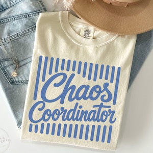 May include: A cream-colored short-sleeve t-shirt featuring the blue text Chaos Coordinator in a script font, centered between vertical blue lines. The shirt is paired with light blue denim jeans, a tan wide-brim hat, and a silver necklace.
