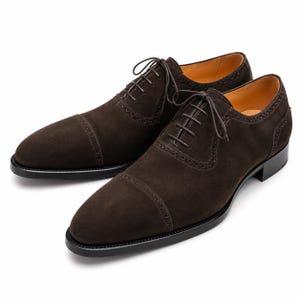 Könnte beinhalten: Ein Paar dunkelbraune Wildleder-Oxford-Schuhe mit einer Zehenkappe, dezenten Brogue-Details entlang der Nähte, dünnen passenden Schnürsenkeln, einem beigen Lederinnenfutter und einer schwarzen Sohle mit einem niedrigen, gestapelten Absatz.