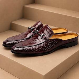 May include: A pair of dark burgundy mule loafers featuring a glossy crocodile-embossed leather texture. Each shoe is adorned with a silver-toned metal horsebit detail across the vamp. The interior lining is a contrasting mustard yellow color.