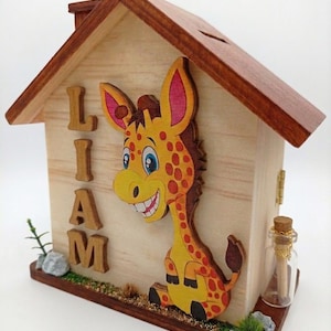 Könnte beinhalten: Eine hölzerne Spardose in Hausform mit einem Ausschnitt einer gelb-orangen Giraffe mit roten Flecken. Der Name LIAM ist in erhabenen Holzbuchstaben auf der linken Seite angebracht. Eine kleine Glasflasche mit Korken steht auf dem Sockel.