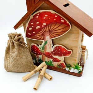 Könnte beinhalten: Eine hausförmige Holzbox mit einem Münzschlitz auf dem Dach, verziert mit einer Illustration von rot-weißen Pilzen. Dazu gehören ein kleiner Jutebeutel, drei mit Schnur gebundene Papierrollen und ein winziges Glasfläschchen mit einem Korken.