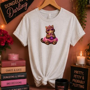 May include: A heathered light grey t-shirt featuring a graphic of a brown tabby cat wearing a pink tiara, pink heart-shaped sunglasses, and a pink tutu while holding a purple potion bottle. The shirt has a crew neckline and short sleeves.