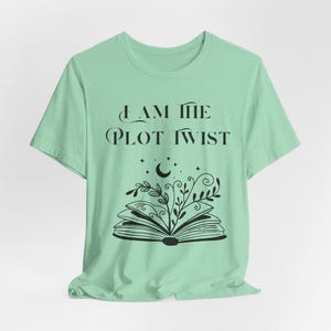 May include: A light green t-shirt with the text "I AM THE PLOT TWIST" in black. Below the text is a black illustration of an open book with a crescent moon, stars, and plant design.