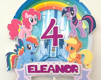 May include: A circular cake topper featuring a rainbow, a castle, and four cartoon ponies. The number 4 is in purple glitter in the center. A light purple banner at the bottom displays the name ELEANOR in purple glitter text on a white background.