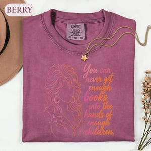 May include: A berry-colored t-shirt with a Comfort Colors label, featuring a line-art illustration of a person with voluminous hair and the text: You can never get enough books into the hands of enough children. A gold star necklace rests on the collar.