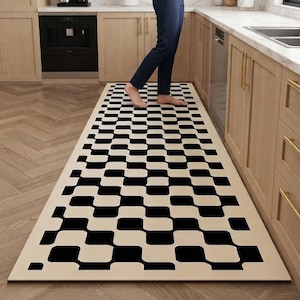 May include: A long, rectangular runner rug featuring a repeating black and cream-coloured pattern of rounded, interlocking squares. The rug is positioned on a wooden floor in a kitchen setting, extending lengthwise between light wood cabinets.