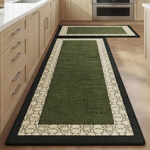 May include: A set of two rectangular kitchen rugs featuring a solid olive green centre, a cream-coloured border with a repeating intricate line pattern, and a solid black outer edge. The rugs are positioned on a light wood floor in a kitchen.