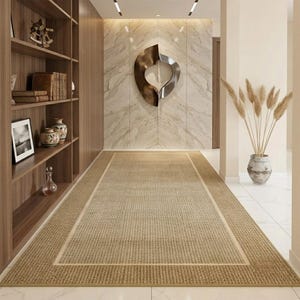May include: A rectangular area rug with a textured, woven tan surface and a lighter beige border. The rug is positioned in a hallway with wood shelving on the left, a marble-patterned wall with a metallic sculpture, and a vase with dried grass.