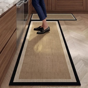 Puede incluir: Una alfombra rectangular de cocina con un centro de color tostado texturizado, un borde interior beige claro y un borde exterior negro sólido. La alfombra está colocada sobre un suelo de madera clara frente a gabinetes de cocina.