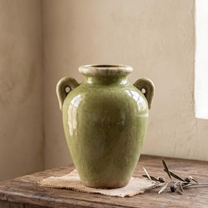 Könnte beinhalten: Eine grüne Keramikvase mit zwei Henkeln, auf einer Jutematte auf einem Holztisch. Die Vase hat eine glänzende Oberfläche und ein leicht gealtertes Aussehen. Ein Olivenzweig mit Oliven liegt auf dem Tisch.