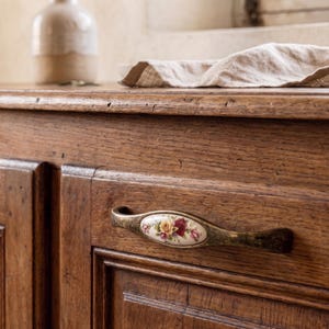 Könnte beinhalten: Nahaufnahme eines Holzschranks mit einem dekorativen Griff. Der Griff hat einen floralen Keramikeinsatz mit roten, gelben und weißen Rosen. Der Schrank ist aus dunkelbraunem Holz gefertigt.