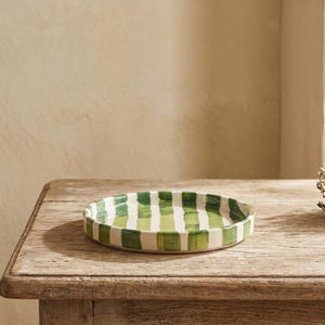Op de afbeelding: Een ronde keramische serveerschaal met een witte basis en verticale groene strepen. De schaal staat op een verweerde houten tafel. Rechts van de schaal ligt een klein bosje gedroogde kruiden.